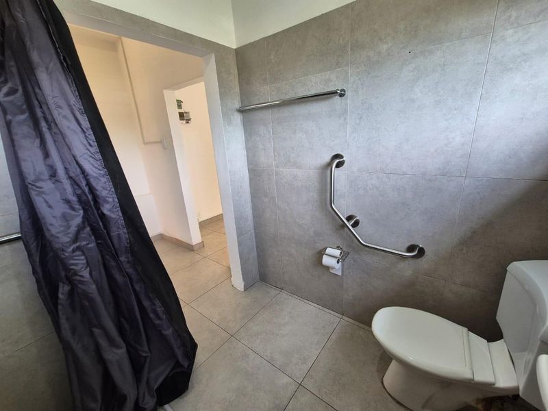 A functional bathroom featuring grey wall and floor tiles, a toilet, and a stainless steel grab rail for accessibility.