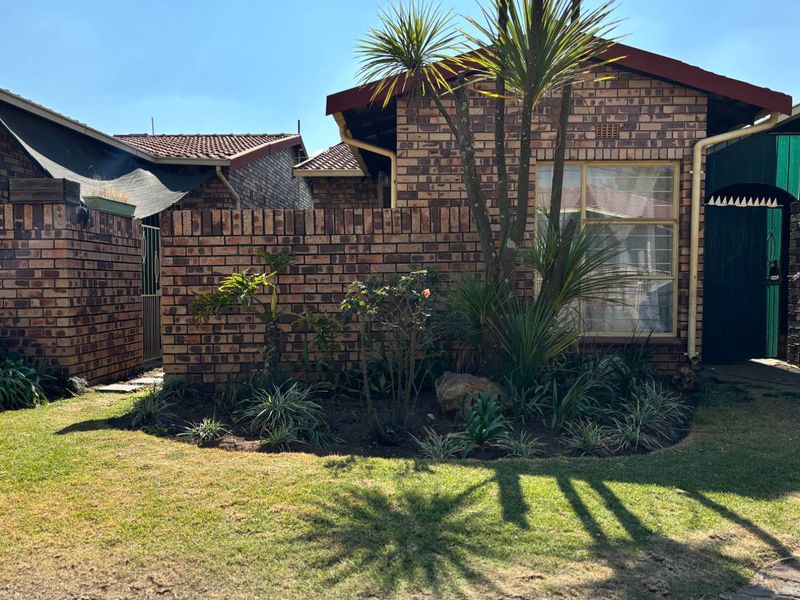 Front elevation of a face brick townhouse featuring a small landscaped garden, security gate, and tiled roof structure.