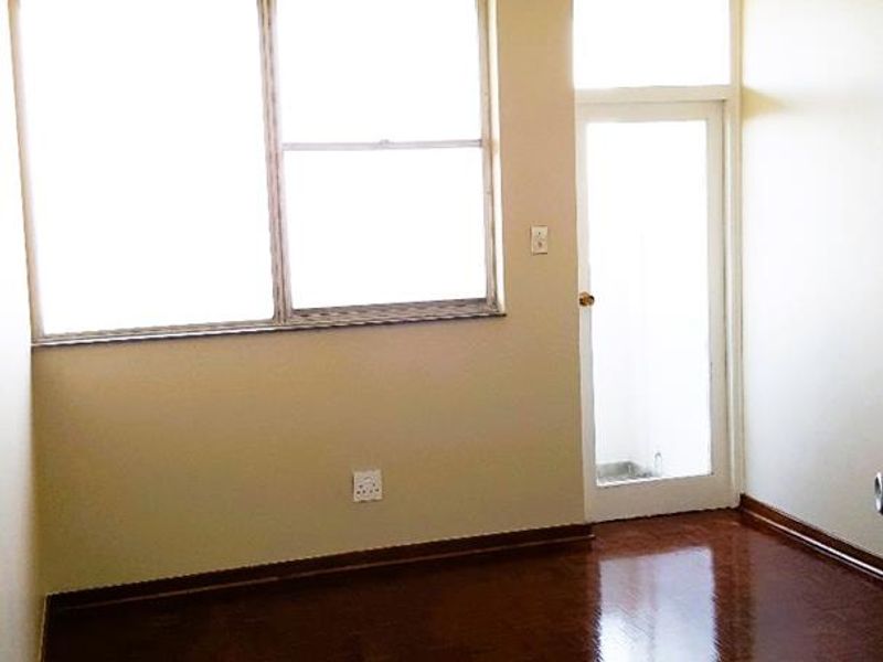 A bright, empty bedroom featuring polished parquet flooring, a large window for natural light, and a glass-paneled door leading to an outdoor area.