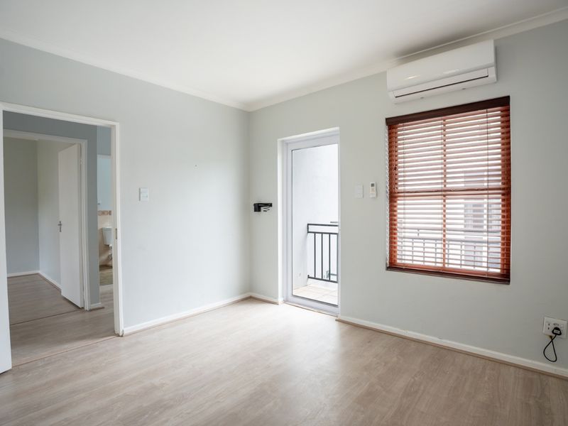 A bright, empty bedroom featuring light laminate flooring, a split-system air conditioner, timber blinds, and direct access to a small balcony.