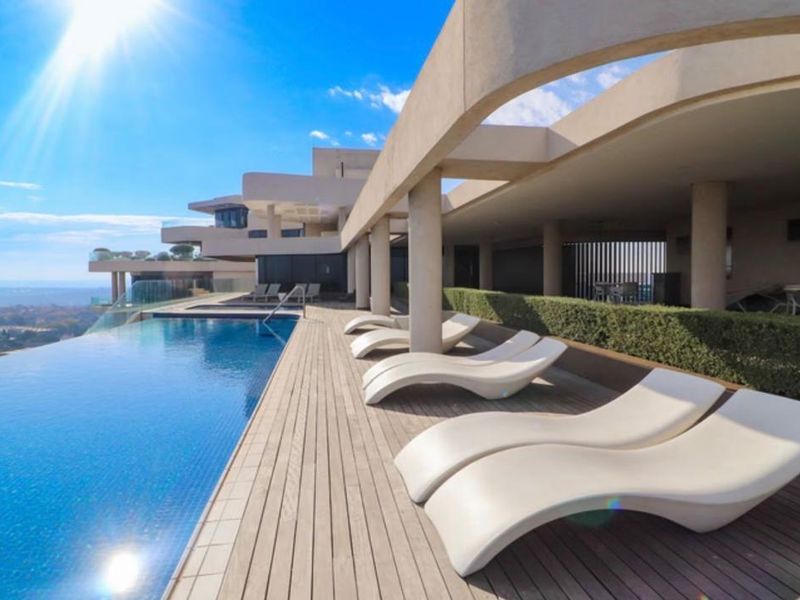 A luxury infinity pool area featuring timber decking, concrete structural columns, and a covered patio space with panoramic views.