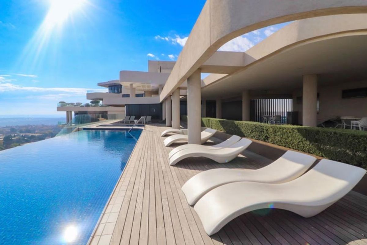 A luxury infinity pool area featuring timber decking, concrete structural columns, and a covered patio space with panoramic views.
