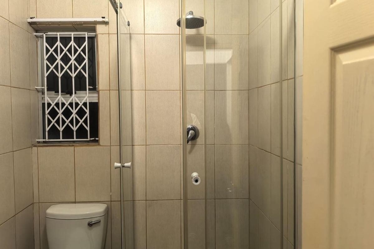 A functional bathroom featuring a glass-enclosed shower with a rain shower head, a toilet, and neutral wall tiling with a security-gated window.