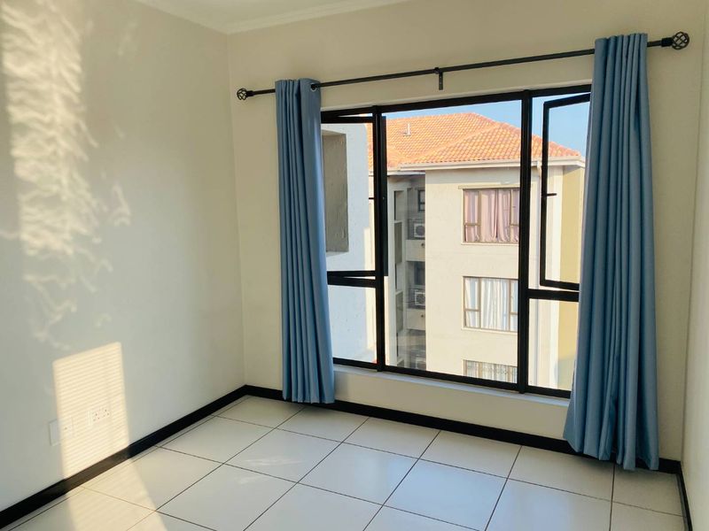 A bright, empty bedroom featuring neutral tiled flooring and large black-framed windows with views of the surrounding apartment complex.