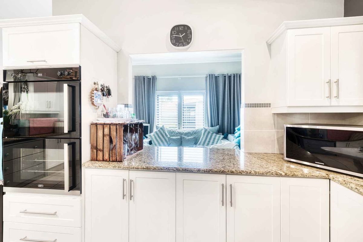 A bright, modern kitchen featuring white cabinetry, granite countertops, and a built-in double oven, with a convenient pass-through window overlooking the living area.