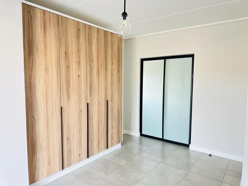 A bright, modern bedroom featuring light wood-grain built-in cupboards, sleek black-framed sliding glass doors, and neutral floor tiles.