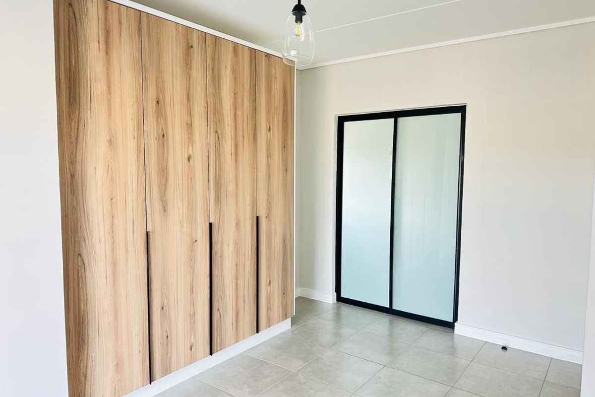 A bright, modern bedroom featuring light wood-grain built-in cupboards, sleek black-framed sliding glass doors, and neutral floor tiles.