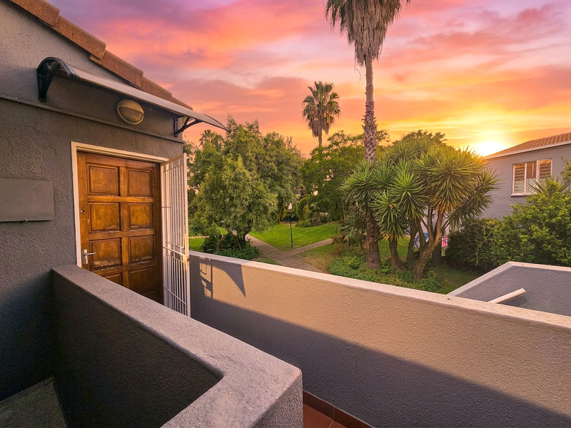 A private balcony area featuring a solid wood entrance door with security gate, overlooking a landscaped garden at sunset.