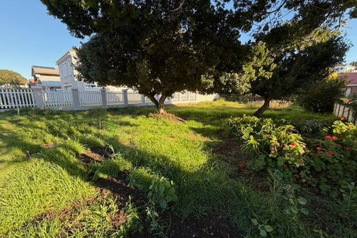 A residential garden area featuring mature trees, established garden beds, and a white picket fence boundary.