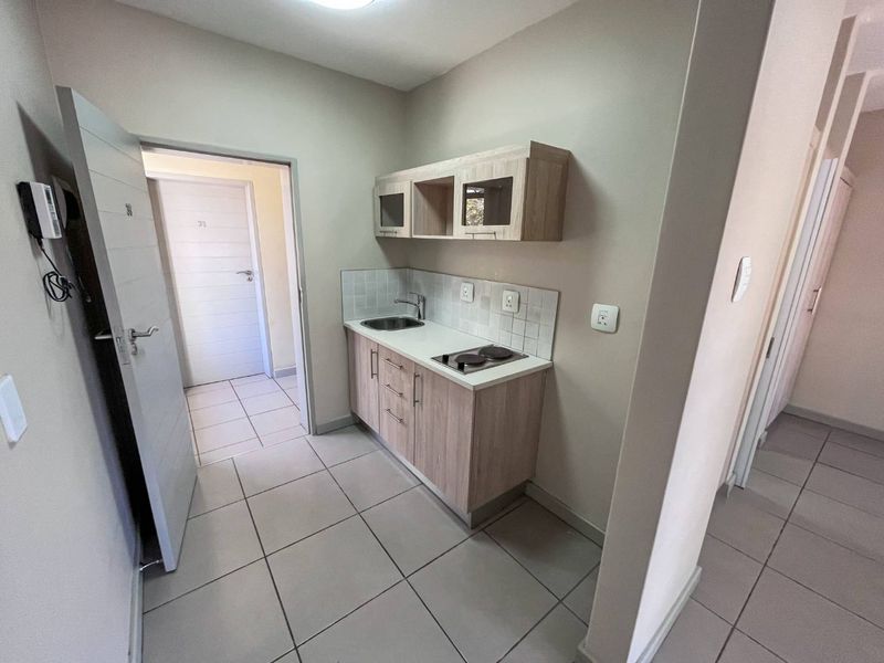 Compact kitchenette featuring light wood-finish cabinetry, a built-in electric hob, stainless steel sink, and neutral tiled flooring in an open-plan layout.