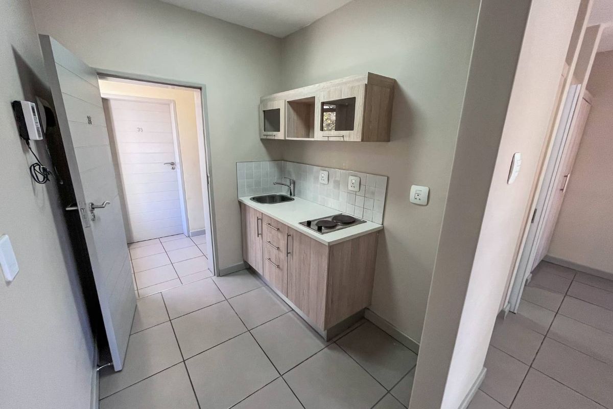 Compact kitchenette featuring light wood-finish cabinetry, a built-in electric hob, stainless steel sink, and neutral tiled flooring in an open-plan layout.