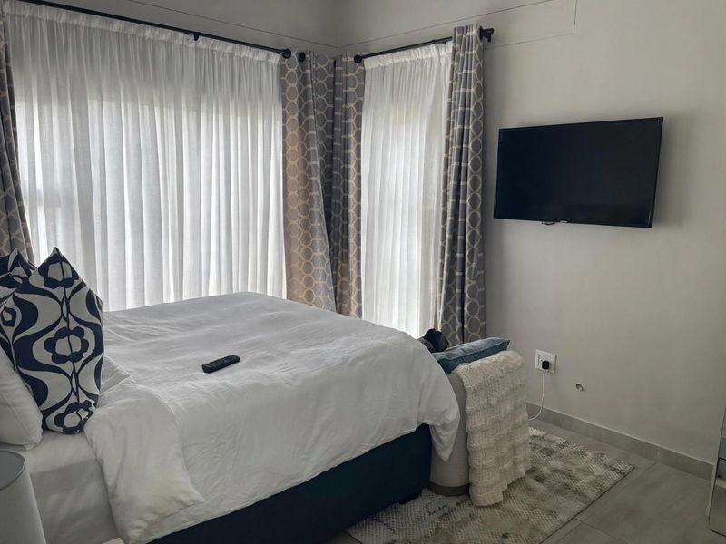 A bedroom featuring tiled flooring, large windows with curtain treatments, and a wall-mounted television.