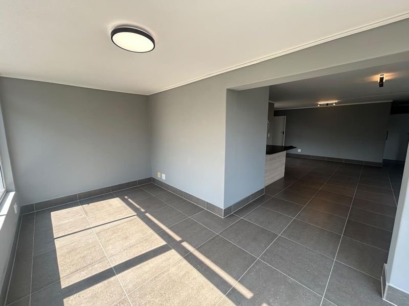 A spacious, empty open-plan living and kitchen area featuring neutral grey walls, modern tiled flooring, and clean architectural lines.