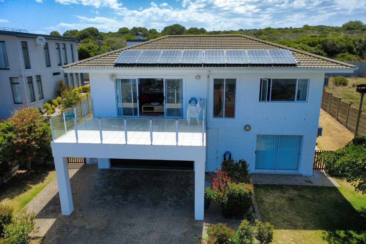 Rear view of a two-story house featuring a raised balcony with glass balustrades, solar panels on the roof, and a paved ground-level area.