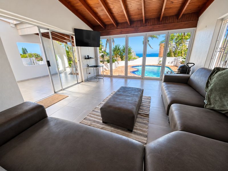 Spacious living area featuring exposed timber beam ceilings, large tiled floors, and expansive sliding glass doors opening to a private swimming pool and ocean view.
