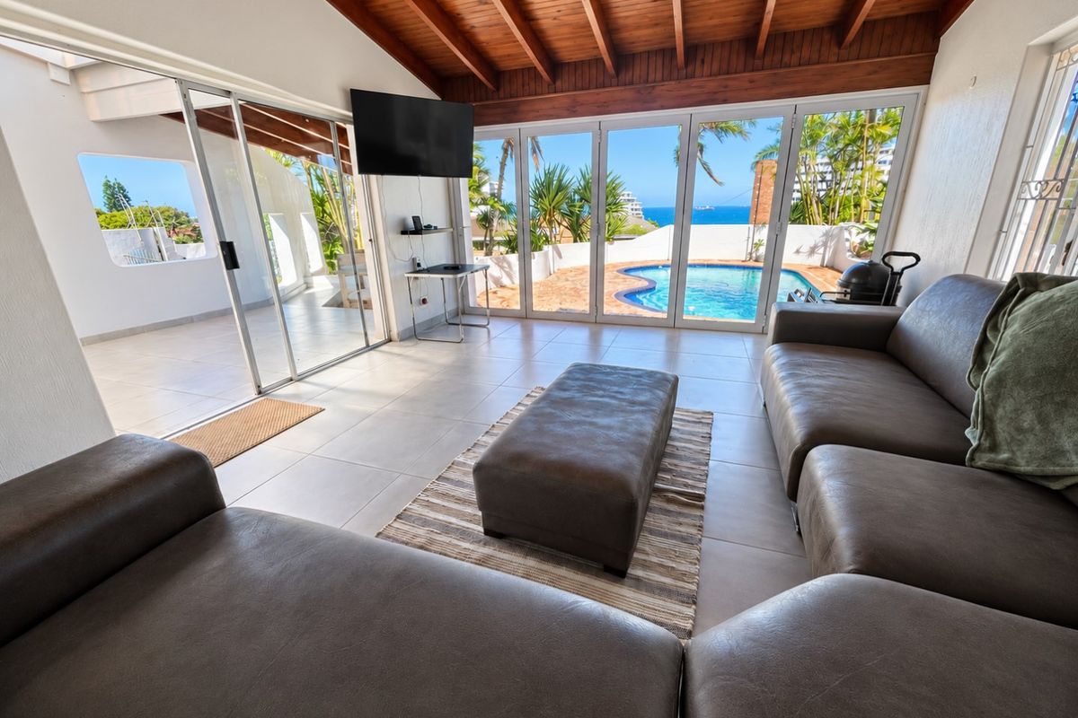 Spacious living area featuring exposed timber beam ceilings, large tiled floors, and expansive sliding glass doors opening to a private swimming pool and ocean view.
