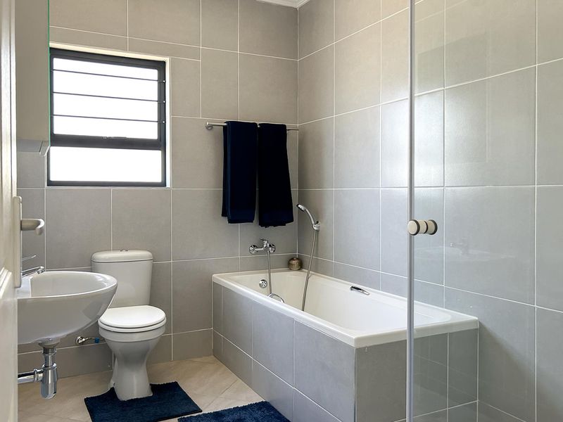 A modern, well-maintained bathroom featuring grey wall tiles, a bathtub with a glass screen, a pedestal basin, and a toilet.