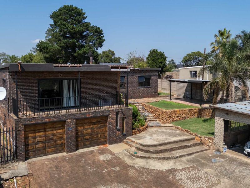 Aerial view of a brick-faced house featuring a double garage, elevated balcony, paved driveway, stone retaining walls, and a separate carport structure.