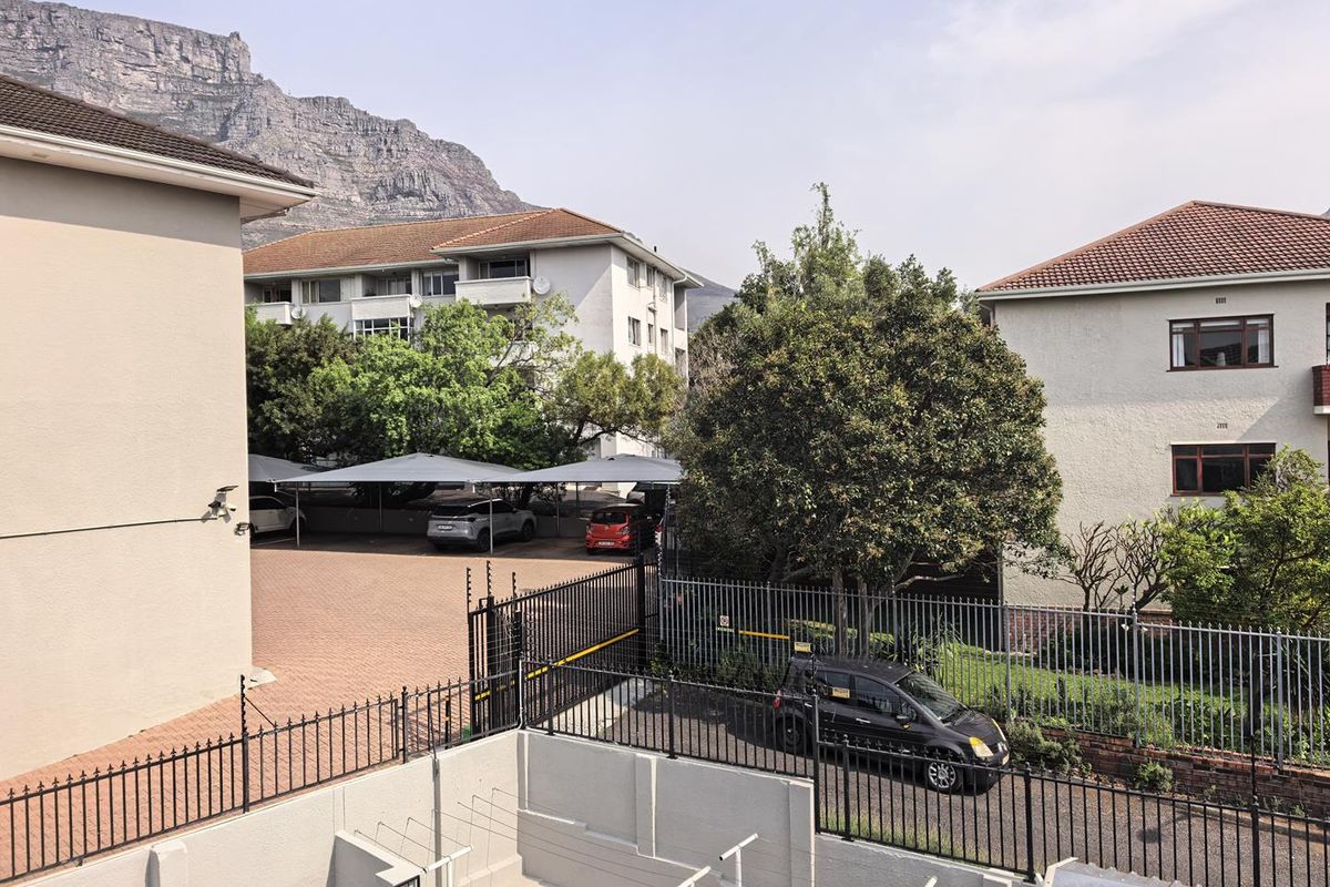 View of the apartment complex exterior, featuring a paved parking area with carports, security fencing, and mountain views in the background.