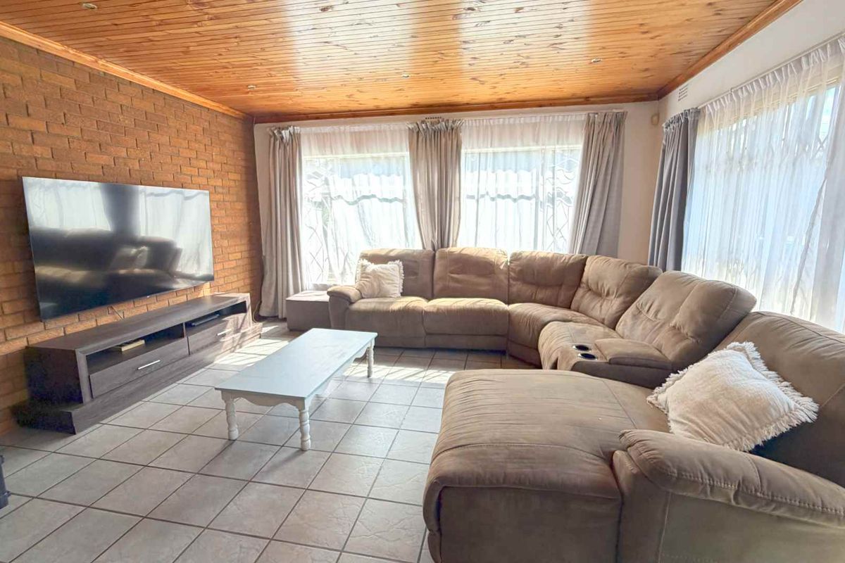 Spacious living room featuring a rustic exposed brick wall, tiled flooring, and a warm timber-paneled ceiling with large windows allowing for ample natural light.