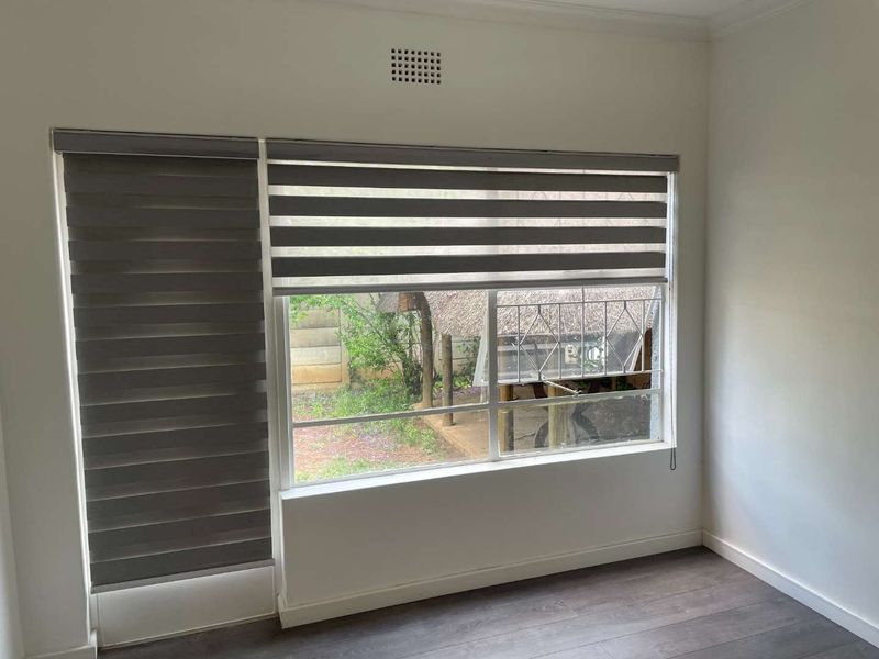 A bright, empty bedroom featuring modern laminate flooring and dual roller blinds, offering a view of the rear garden area.