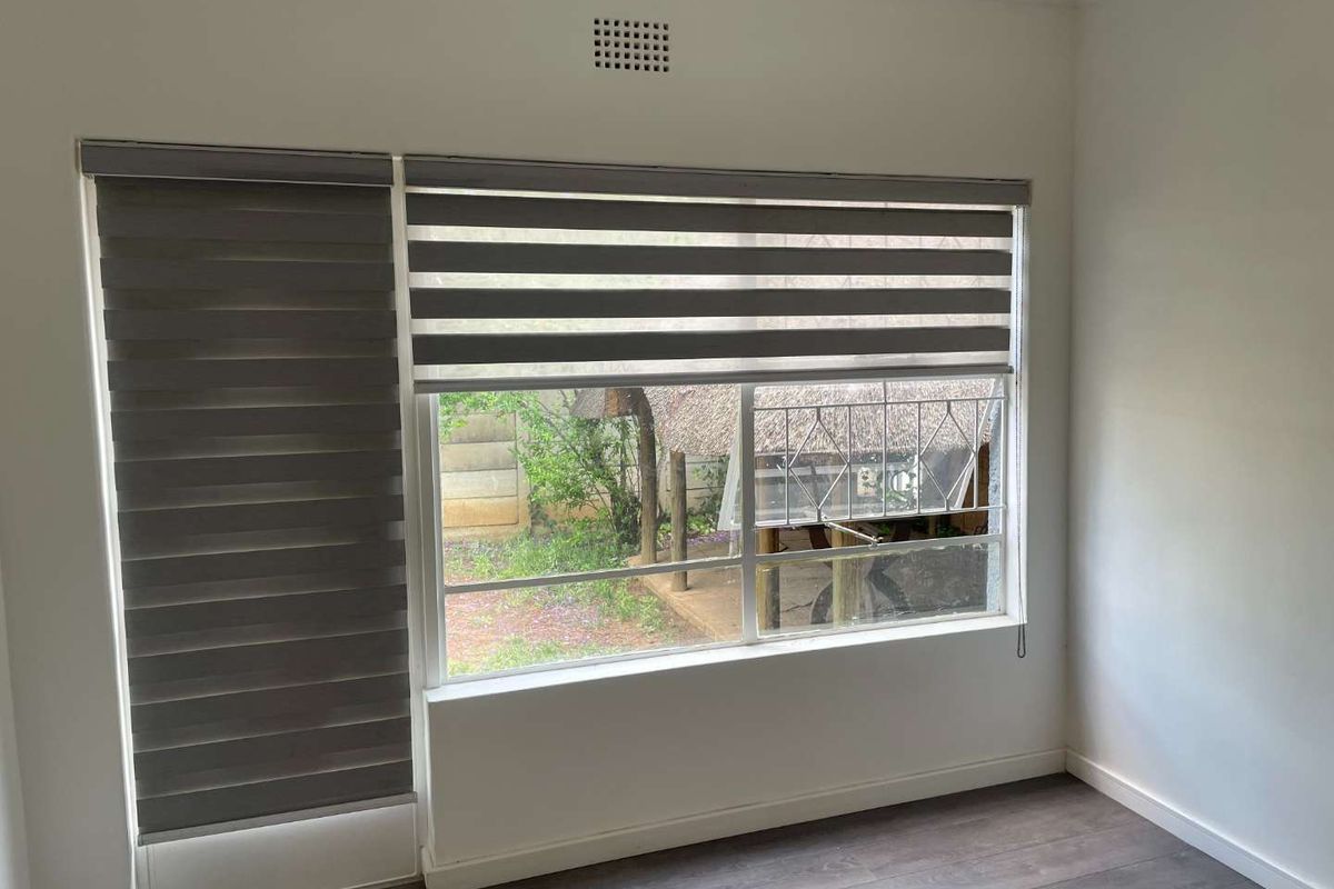 A bright, empty bedroom featuring modern laminate flooring and dual roller blinds, offering a view of the rear garden area.
