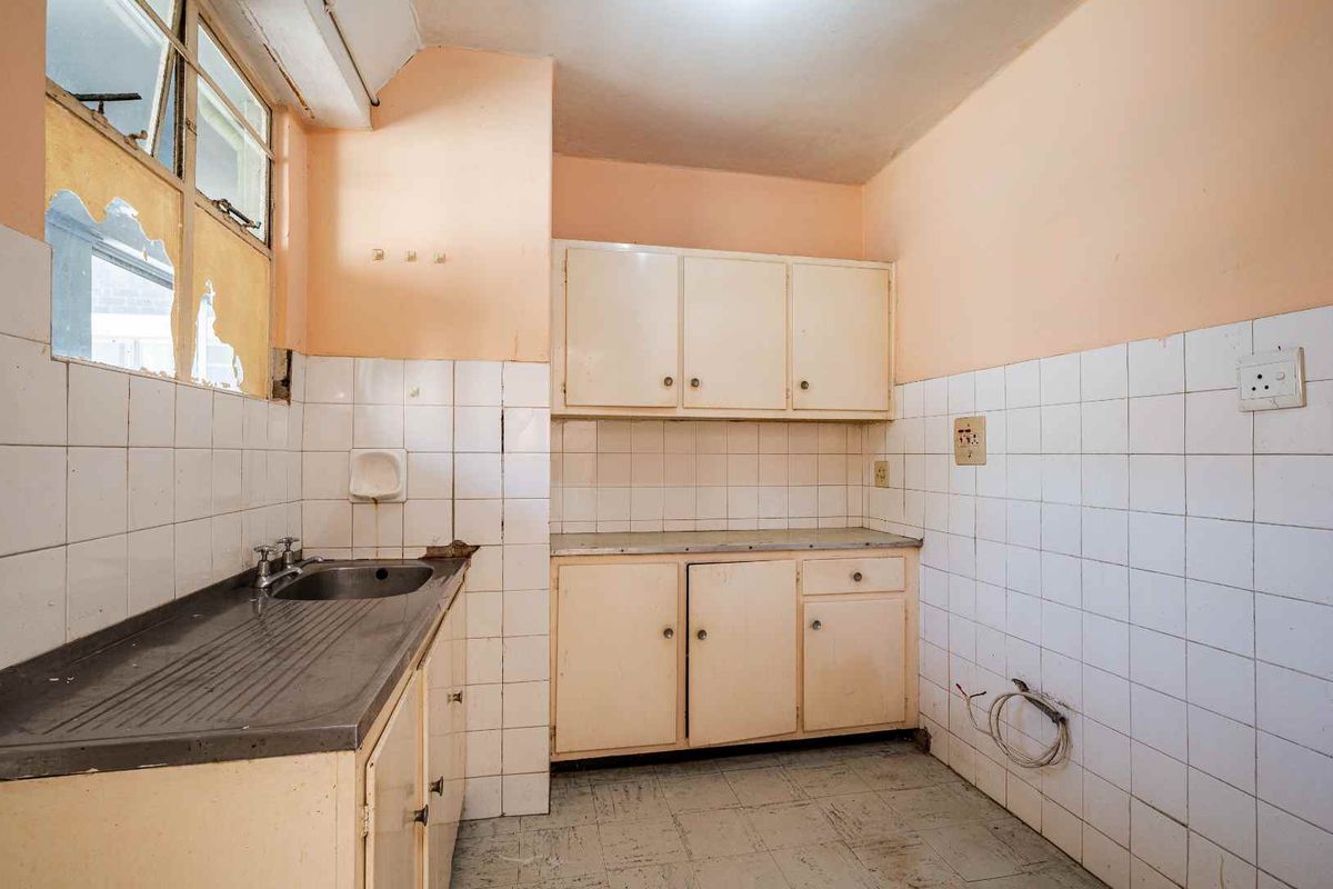 A dated, empty kitchen featuring original cabinetry, a stainless steel sink, and tiled splashbacks, requiring significant renovation and repair to the window and walls.