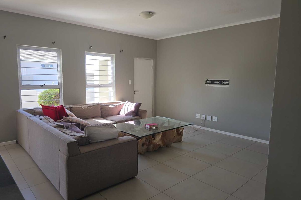 A spacious living room featuring neutral tiled flooring, large windows with security bars, and a wall-mounted TV bracket.