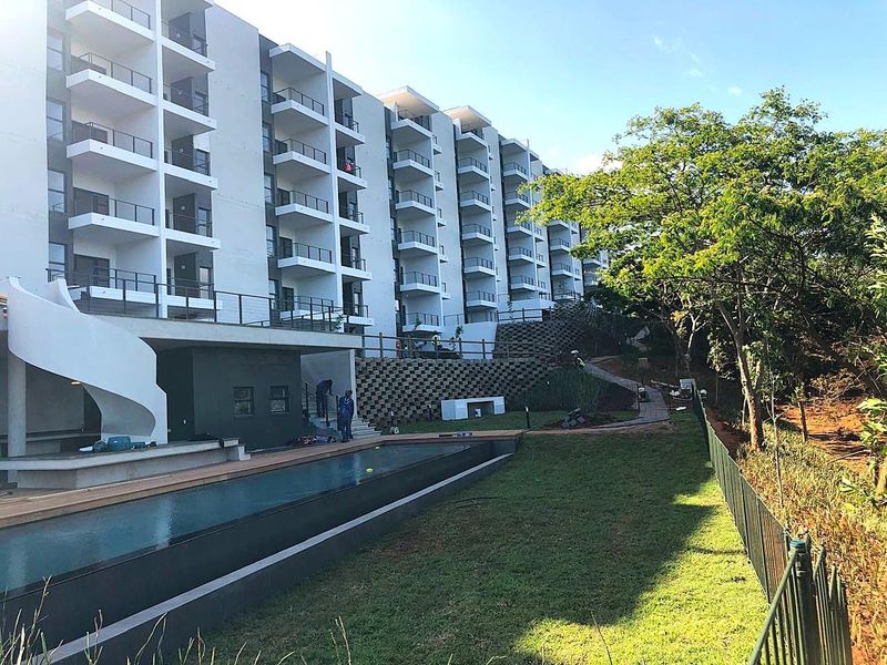 Exterior view of a modern apartment complex featuring a swimming pool, wooden decking, landscaped garden areas, and multiple private balconies.