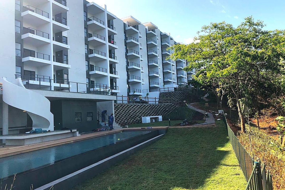 Exterior view of a modern apartment complex featuring a swimming pool, wooden decking, landscaped garden areas, and multiple private balconies.