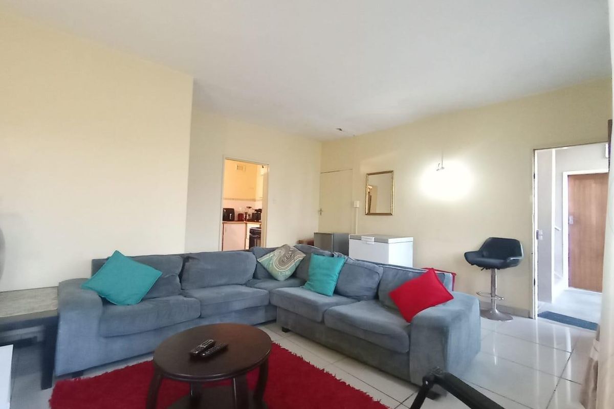 A spacious living room featuring neutral walls and tiled flooring, with direct access to the kitchen and hallway areas.