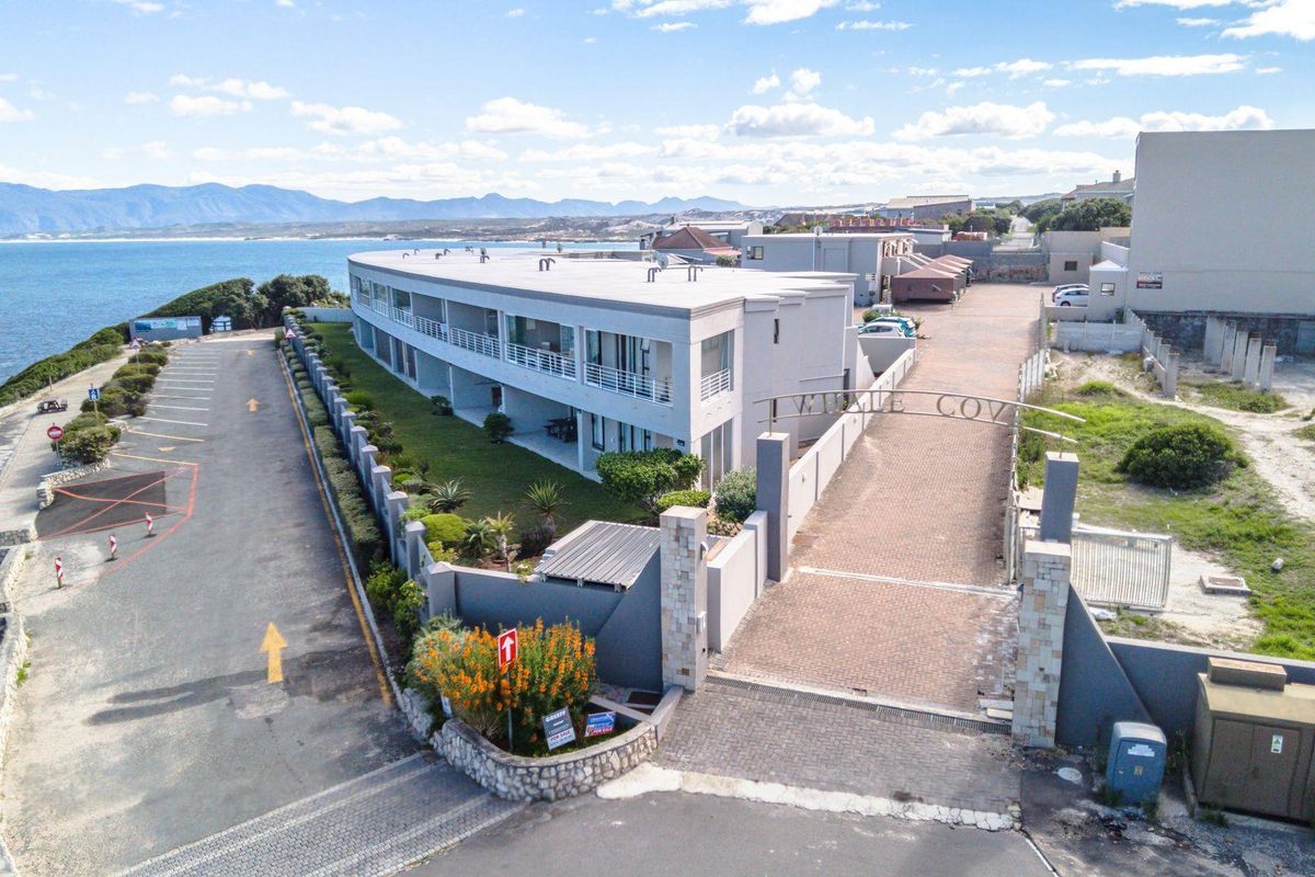 Aerial view of the apartment complex entrance, featuring paved driveway, perimeter wall, and coastal proximity.