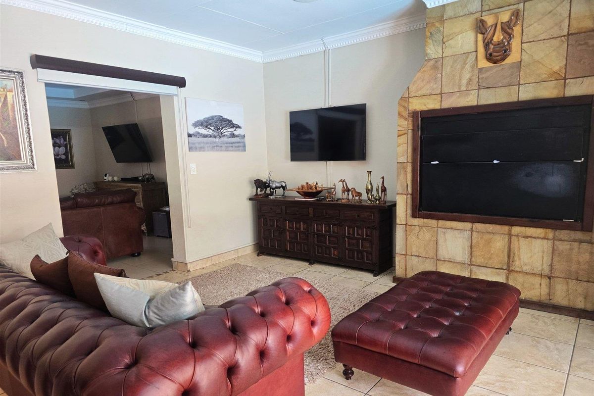 A living room featuring a prominent stone-clad fireplace, tiled flooring, and crown molding, with an open-plan layout leading into an adjacent room.