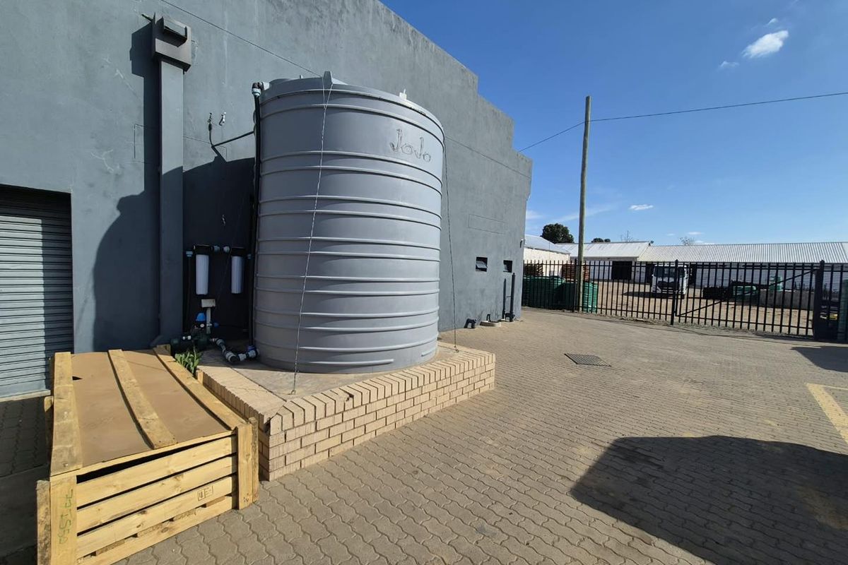 Rear exterior view of the business premises featuring a large water storage tank with filtration system, paved yard, and secure metal fencing.