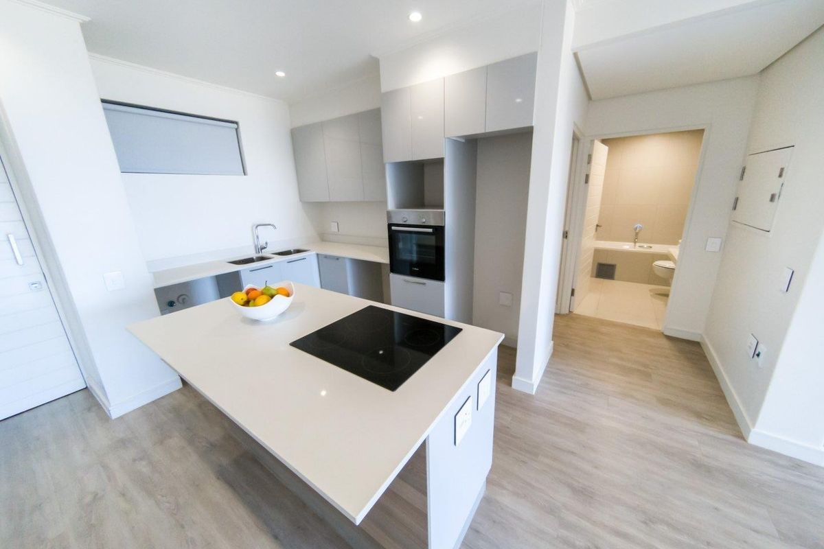 Modern kitchen featuring a central island with an integrated electric hob, built-in oven, sleek cabinetry, and light-toned laminate flooring, with an adjacent bathroom visible.