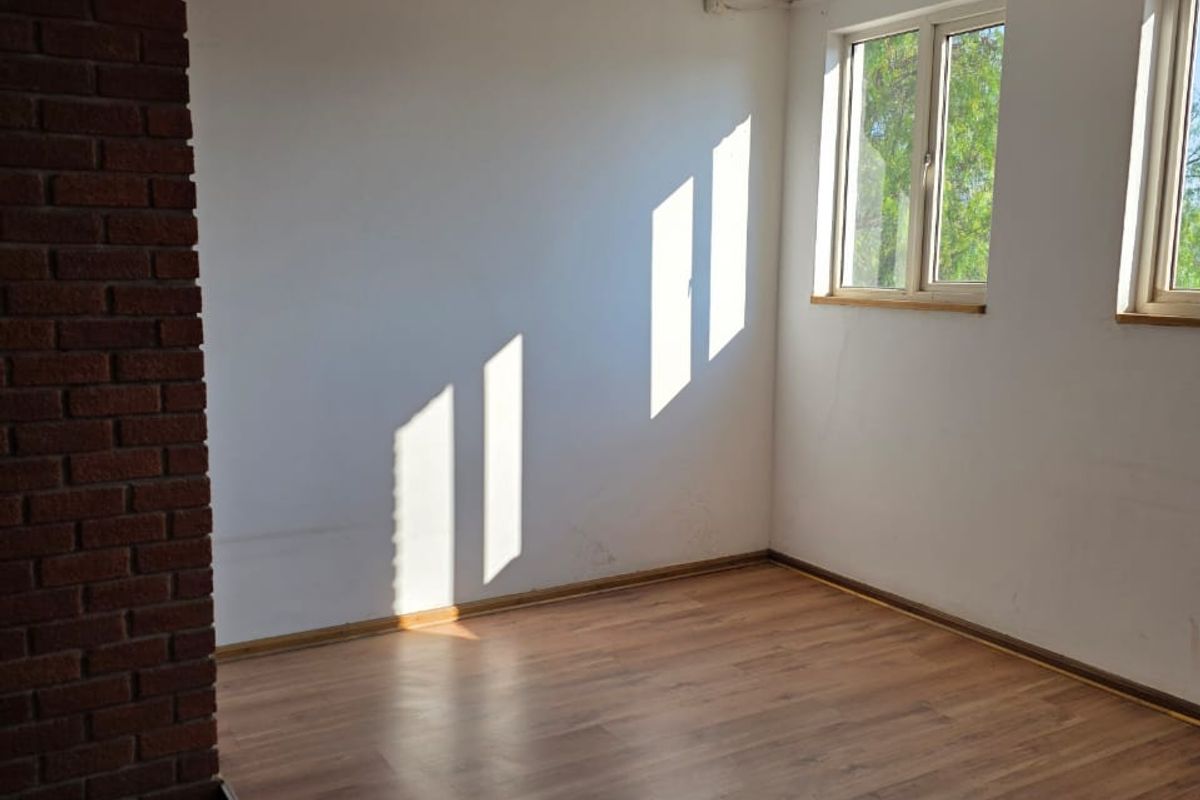 An empty, sunlit room featuring laminate flooring, an exposed brick pillar, and dual windows, offering a blank canvas for industrial or office space customization.