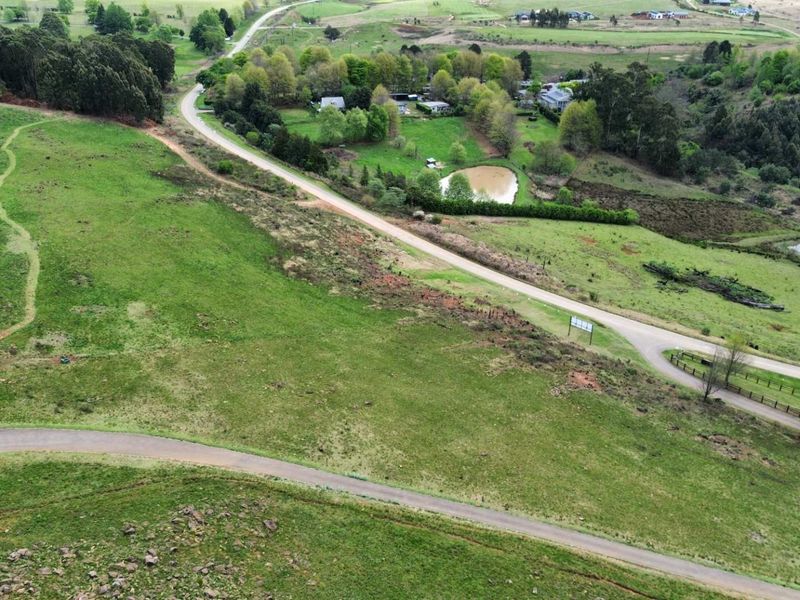 An aerial view of a vacant residential land plot featuring natural grass terrain, paved access roads, and surrounding rural landscape.