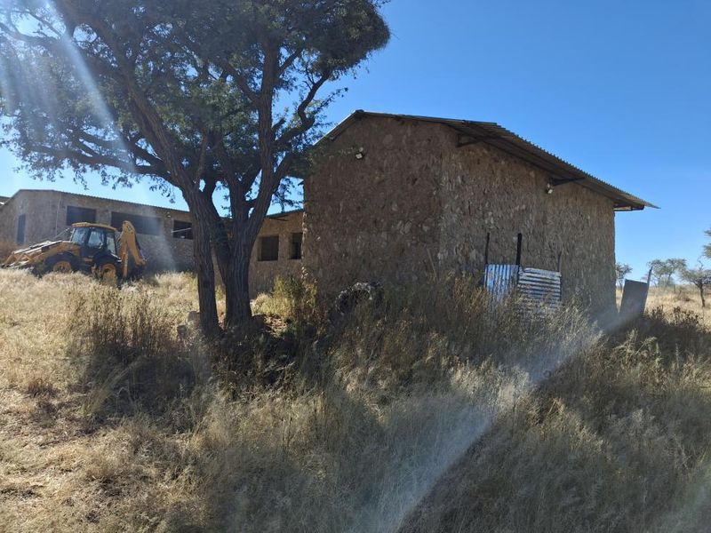Exterior view of a stone-walled structure with a corrugated metal roof, situated on a rural small holding with surrounding natural vegetation.