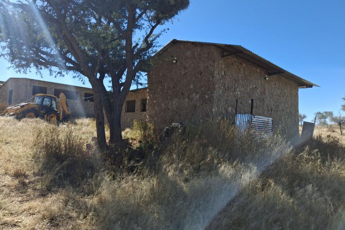 Exterior view of a stone-walled structure with a corrugated metal roof, situated on a rural small holding with surrounding natural vegetation.