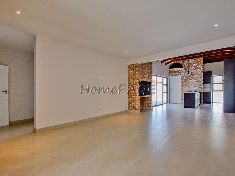 Spacious open-plan living and kitchen area featuring exposed brickwork, timber ceiling beams, tiled floors, and a built-in fireplace.