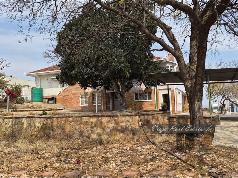 Rear view of the property featuring a brick facade, stone retaining wall, carport, and established trees in a spacious garden setting.