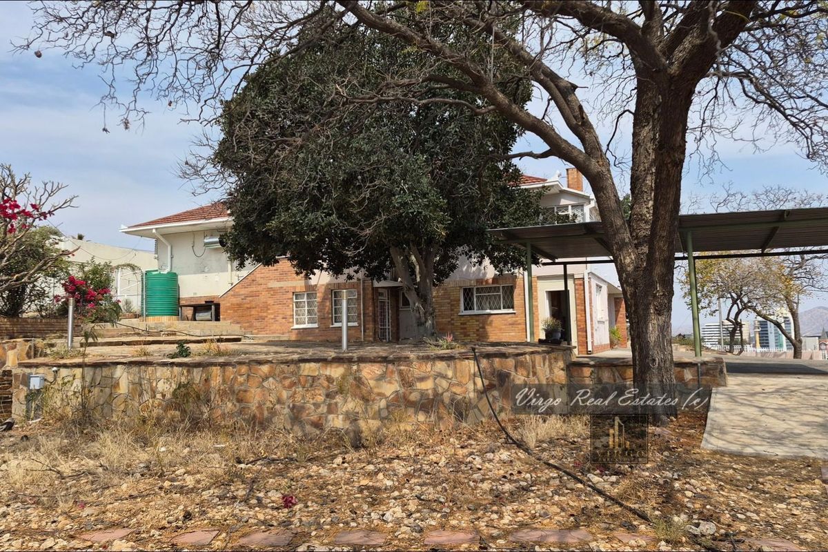 Rear view of the property featuring a brick facade, stone retaining wall, carport, and established trees in a spacious garden setting.
