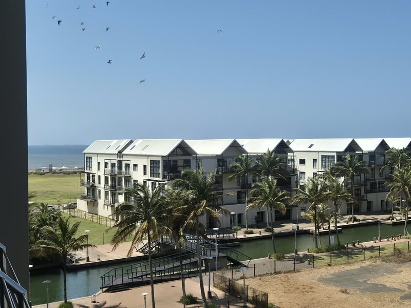 Elevated view overlooking a canal, pedestrian bridge, and landscaped palm trees with a multi-story apartment complex and distant ocean horizon.