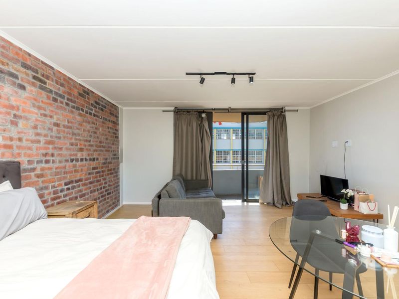 Open plan living and bedroom area featuring an exposed brick feature wall, laminate flooring, and sliding glass doors leading to a balcony.