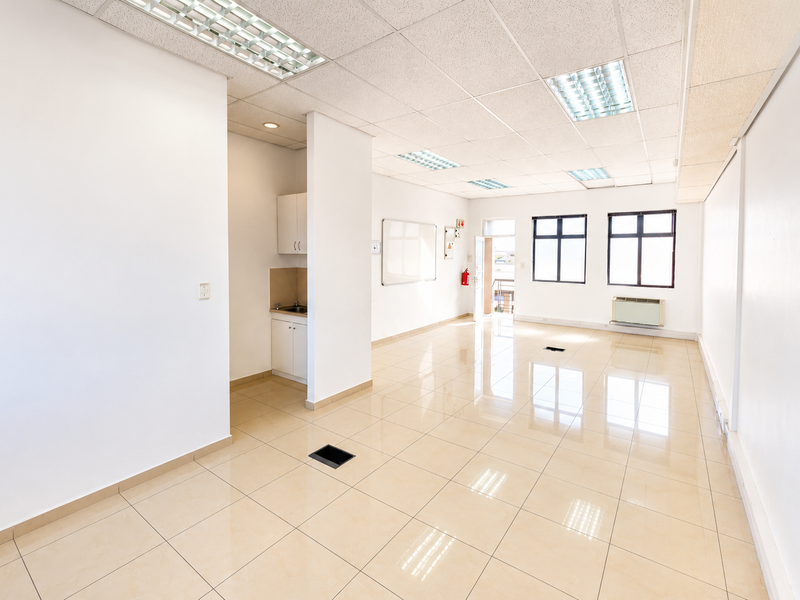 A bright, open-plan office space featuring tiled flooring, a suspended ceiling with recessed lighting, a small kitchenette, and an integrated air conditioning unit.