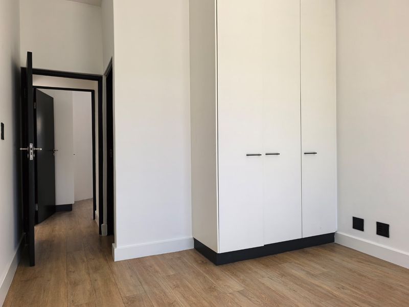 A clean, modern bedroom featuring light wood-look laminate flooring, crisp white walls, and sleek built-in cupboards with contrasting black base detailing.