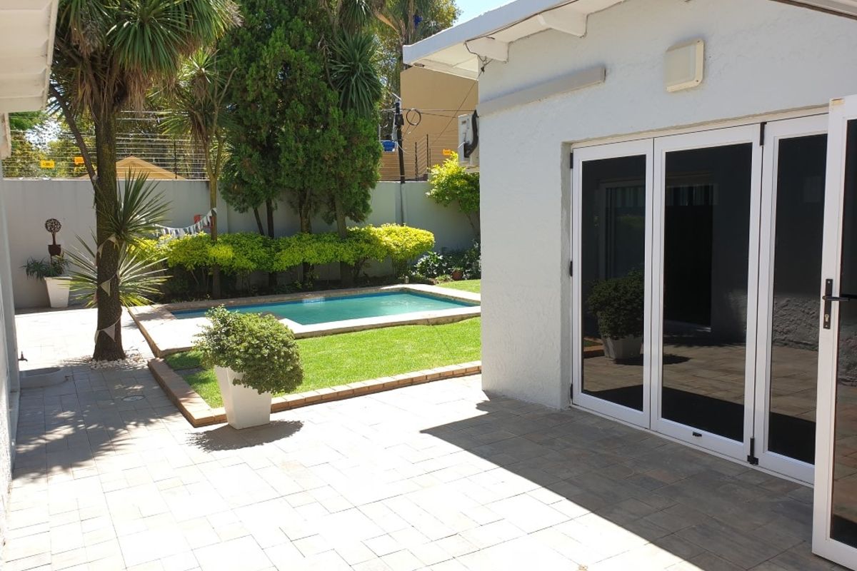 A well-maintained rear garden featuring a paved patio area, a swimming pool, and established landscaping, accessible via modern glass folding doors.