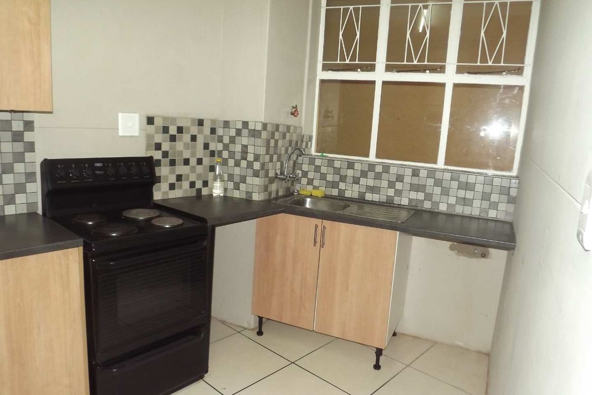 Compact kitchen featuring light wood-finish cabinetry, mosaic tile backsplash, dark countertops, and a functional freestanding stove with security-barred window.