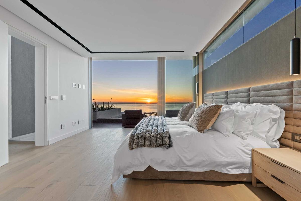 Modern luxury bedroom featuring hardwood floors, an upholstered headboard, and floor-to-ceiling glass doors opening to a sunset ocean view.