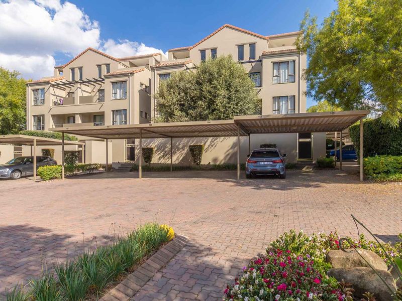 Exterior view of a multi-story apartment complex featuring covered parking bays, a paved driveway, and established garden landscaping.